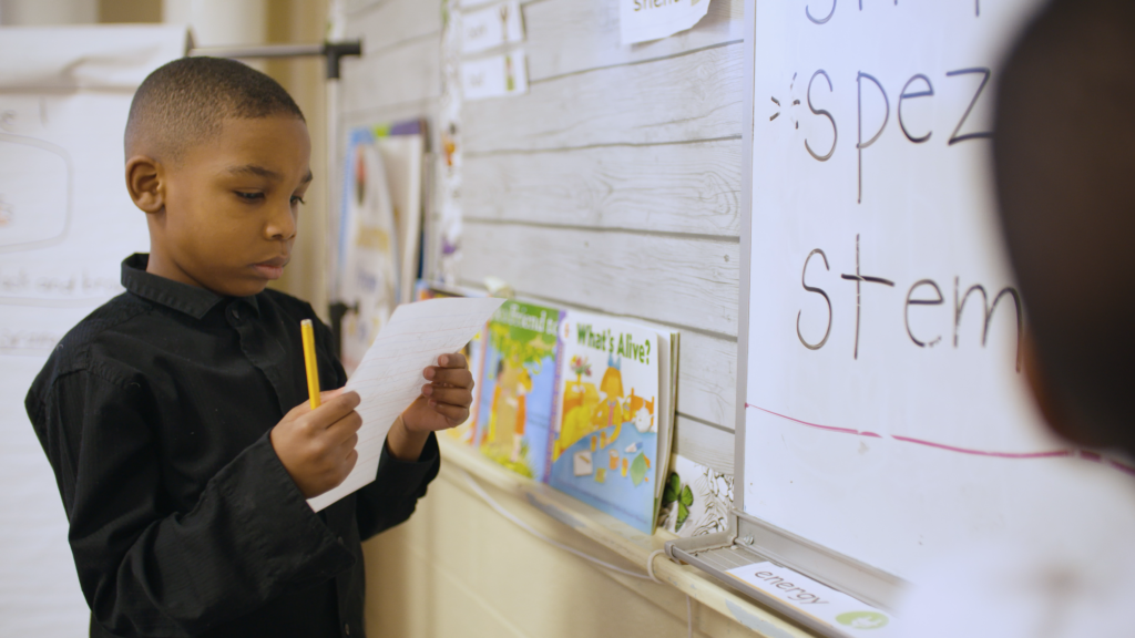 Fred, a featured student in The Right to Read film stands at a classroom board holding a piece of paper he is reading.